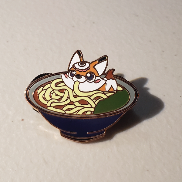 A picture of an enamel pin featuring a fox eating kitsune udon
